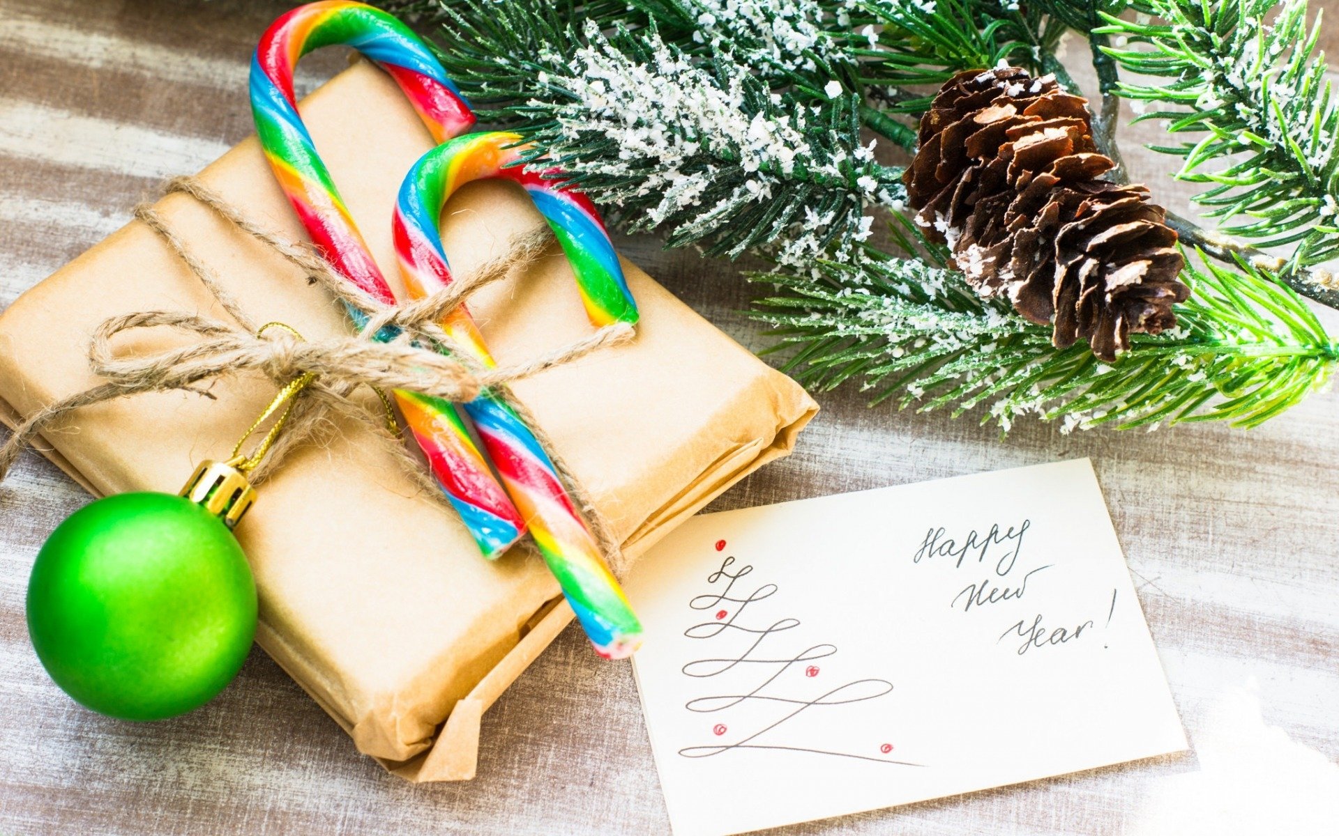 HD holiday desktop wallpaper featuring candy canes, a wrapped gift, green ornament, pinecone, snowy pine branches, and a handwritten Happy New Year note.