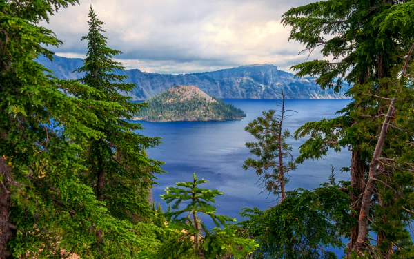 HD desktop wallpaper of Crater Lake showcases serene blue waters surrounded by lush forest and tree-covered landscape, under a cloudy sky, creating a breathtaking natural scene.