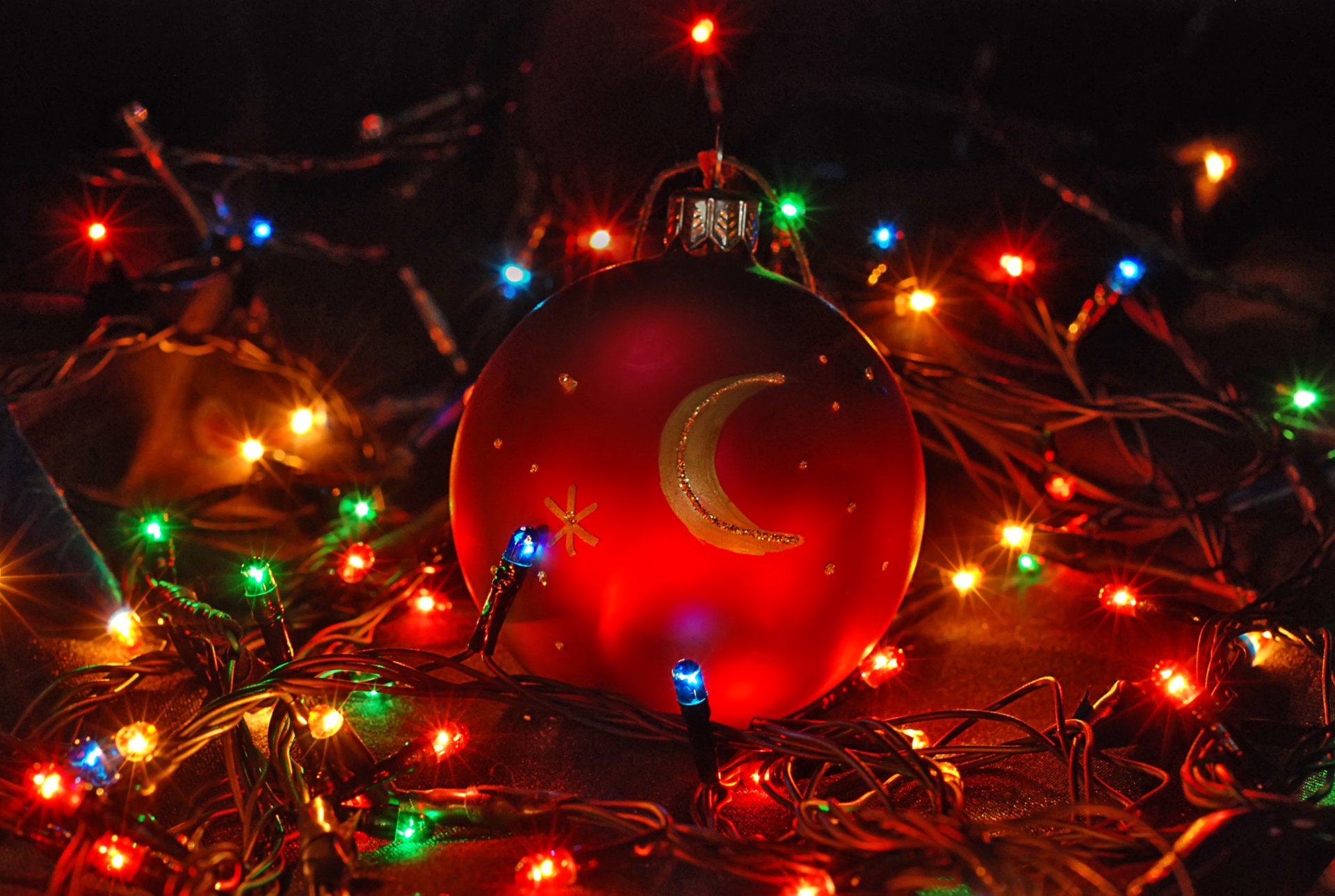 Festive HD desktop wallpaper of a red Christmas ornament with a crescent moon design, surrounded by colorful holiday lights.