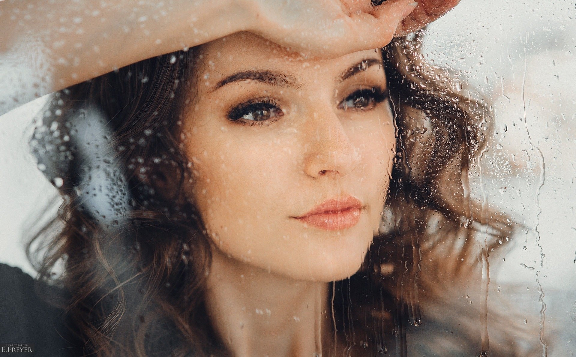 A high-definition wallpaper featuring a brunette model with brown eyes, gazing pensively through raindrops on a window.