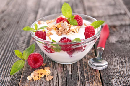 A vibrant 4K Ultra HD image of a glass bowl filled with yogurt, fresh raspberries, muesli, and mint leaves on a rustic wooden surface with a spoon nearby.