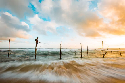 HD PC desktop wallpaper: fisherman perched on a stilt in the ocean, fishing as waves churn beneath a dramatic cloud-filled sky — evocative sea photography highlighting people at work.