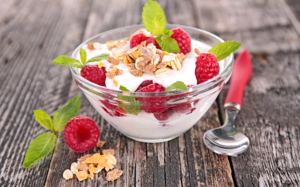 A vibrant 4K Ultra HD image of a glass bowl filled with yogurt, fresh raspberries, muesli, and mint leaves on a rustic wooden surface with a spoon nearby.