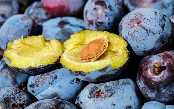Close-up of fresh plums, featuring one halved with its pit visible, captured in sharp detail for a vibrant 4K Ultra HD PC desktop wallpaper background.