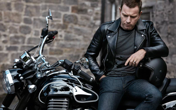 Scottish actor Ewan McGregor in a black leather jacket sitting beside a shiny motorcycle, set against a stone wall in this HD desktop wallpaper.