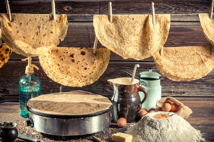 A 4K Ultra HD still life of crêpes drying on a string above a wooden table with eggs, flour, a crêpe maker, and rustic kitchen jars on the floor.