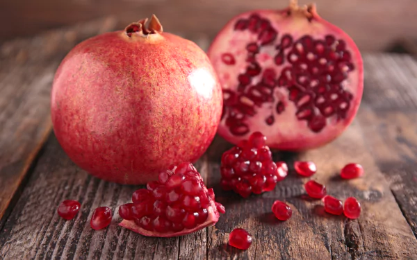 Close-up of whole and halved pomegranate with scattered seeds on wooden surface, captured in vibrant detail for a 4K Ultra HD desktop wallpaper showcasing fresh fruit.