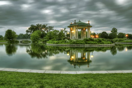 A serene park scene with a man-made gazebo on a small island, surrounded by trees and calm river water reflecting the cloudy sky, captured in HD for desktop wallpaper.