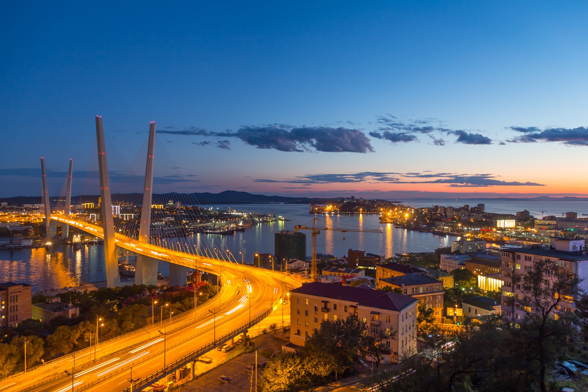 Night view of Vladivostok’s Zolotoy Bridge spanning the coast with city lights reflecting on the sea under a twilight sky on the horizon.