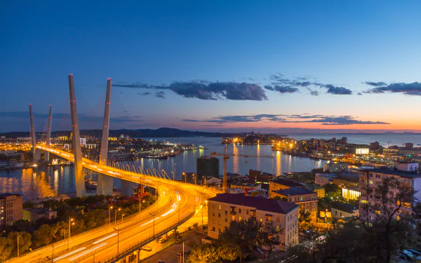 Night view of Vladivostok’s Zolotoy Bridge spanning the coast with city lights reflecting on the sea under a twilight sky on the horizon.