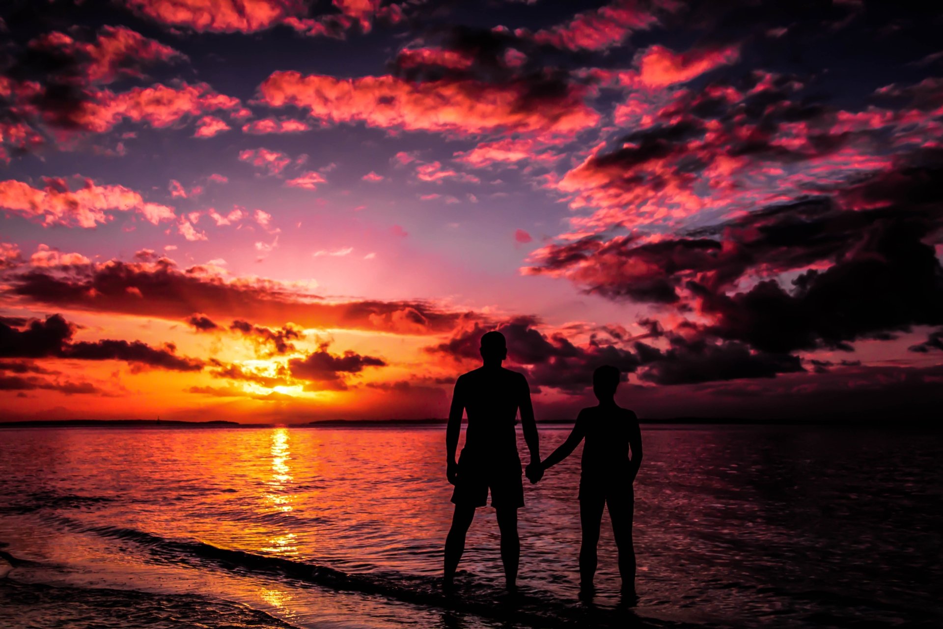 Silhouette of a couple holding hands against a stunning sunset over the ocean, with vibrant colors in the sky and clouds. The scene captures love and tranquility in Australia. HD wallpaper and background.
