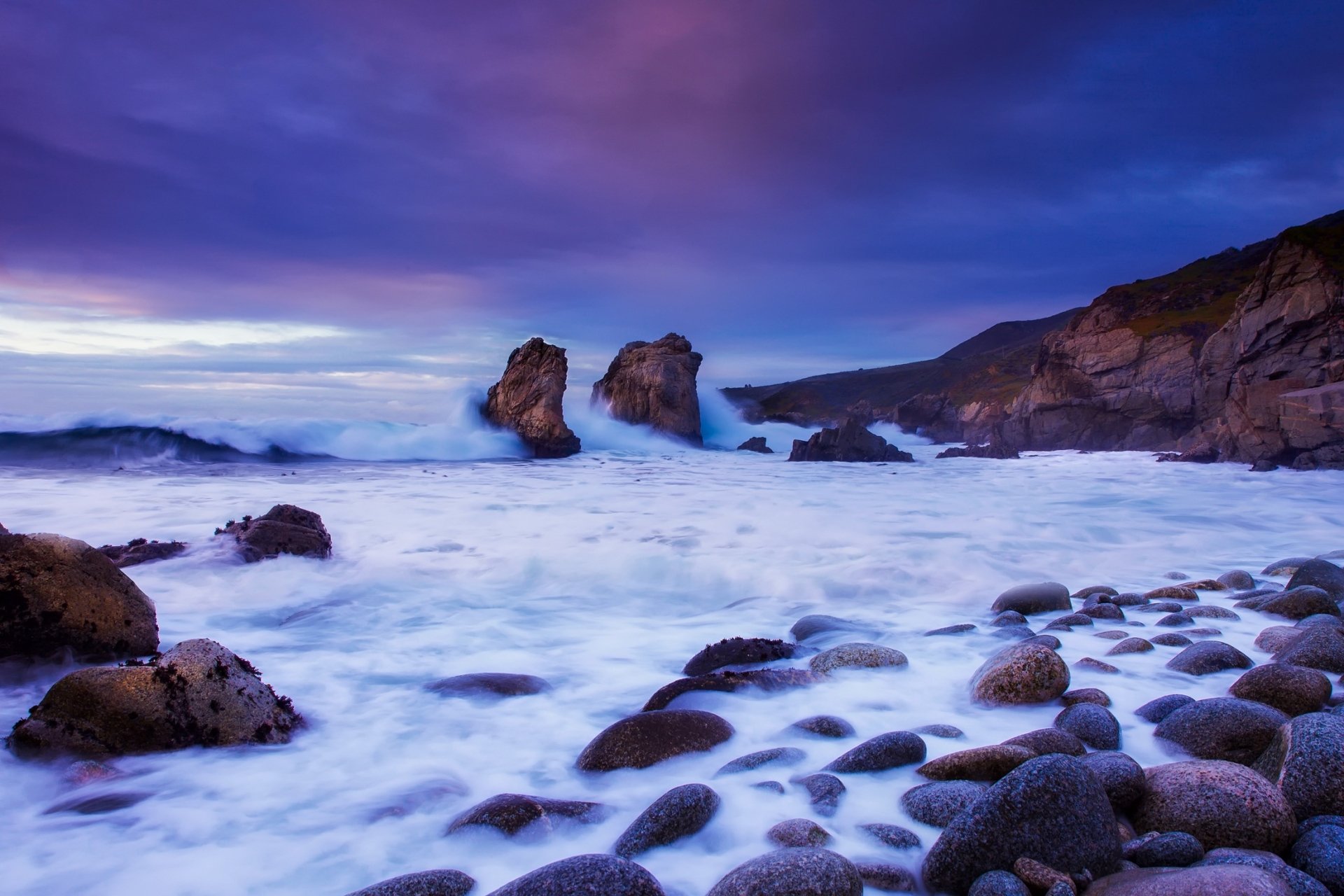 HD PC desktop wallpaper: California coastline nature scene at dusk, ocean waves washing a pebbled seashore with rocky outcrops under a purple-blue sky.