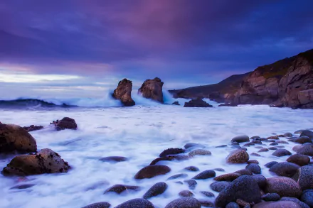 HD PC desktop wallpaper: California coastline nature scene at dusk, ocean waves washing a pebbled seashore with rocky outcrops under a purple-blue sky.