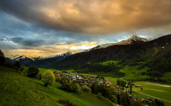 A stunning 4K Ultra HD landscape of a Swiss mountain valley with a small man-made village nestled among green hills under a dramatic cloudy sky.