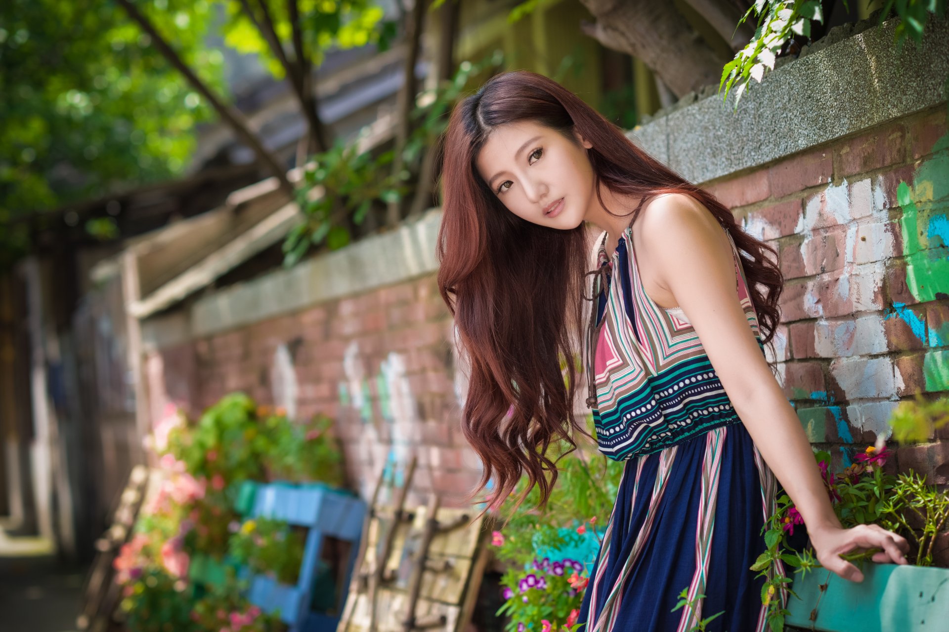 An Asian woman with brown eyes and long brunette hair wears a patterned summer dress, posing outdoors with a blurred garden background and a brick wall, creating a serene, high-definition desktop wallpaper.