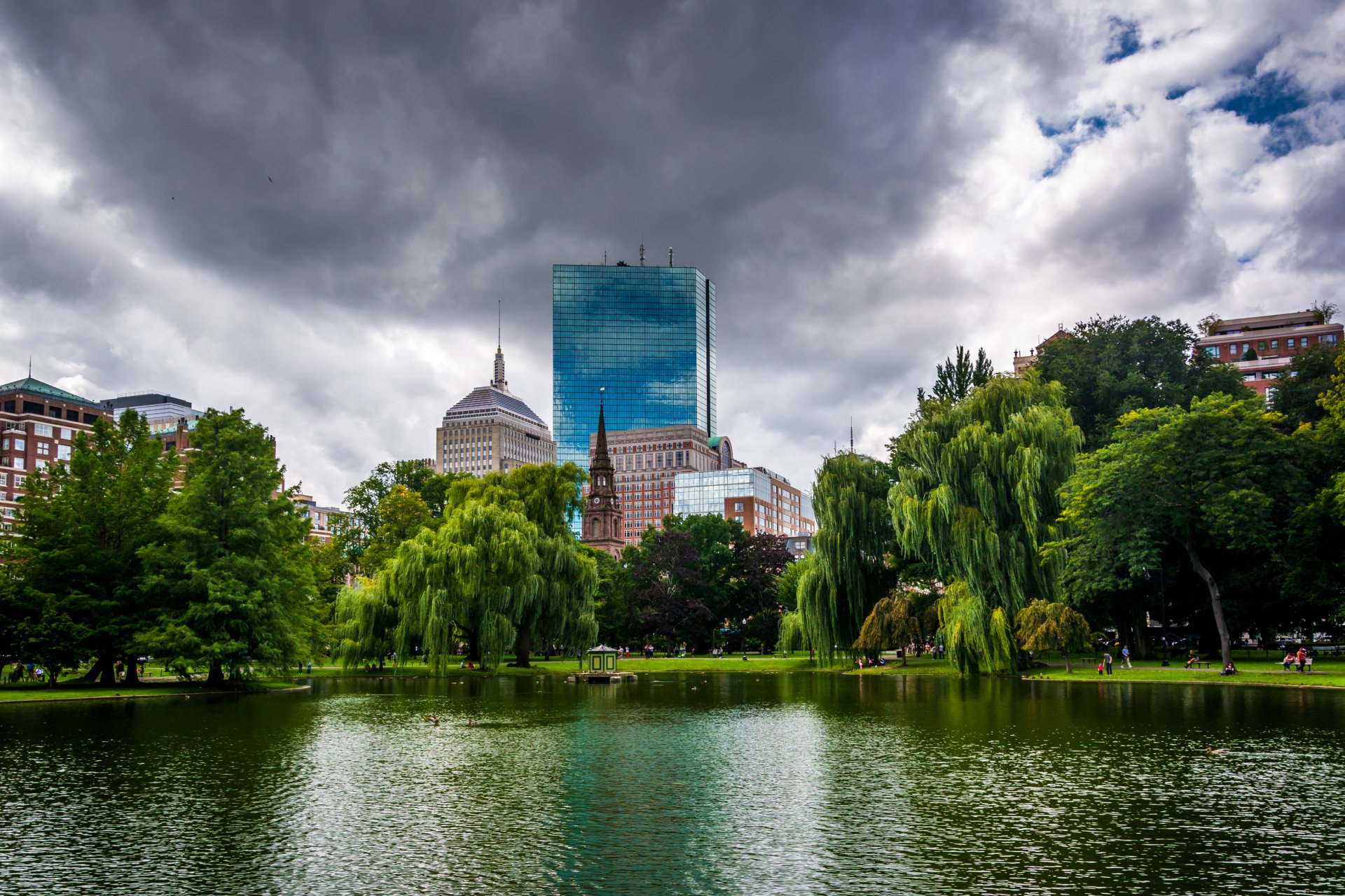 4K Ultra HD desktop wallpaper: Boston park beside the Charles River, willow trees and green lawn with man-made skyline of glass and brick buildings under moody clouds.