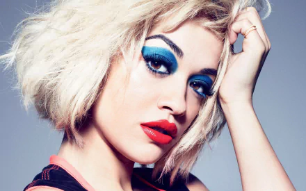 A striking HD desktop wallpaper featuring a close-up of a blonde singer with short hair and bold makeup. Her brown eyes are accented with vibrant blue eyeshadow, and her lips are painted red.
