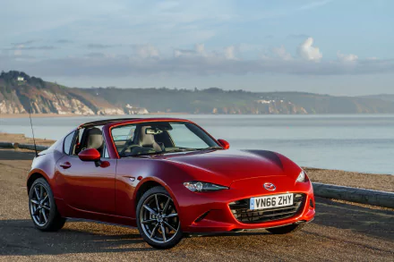 A red Mazda MX-5 sports car parked by a coastal road with cliffs and calm sea under a partly cloudy sky, captured in 4K Ultra HD for PC desktop wallpaper.