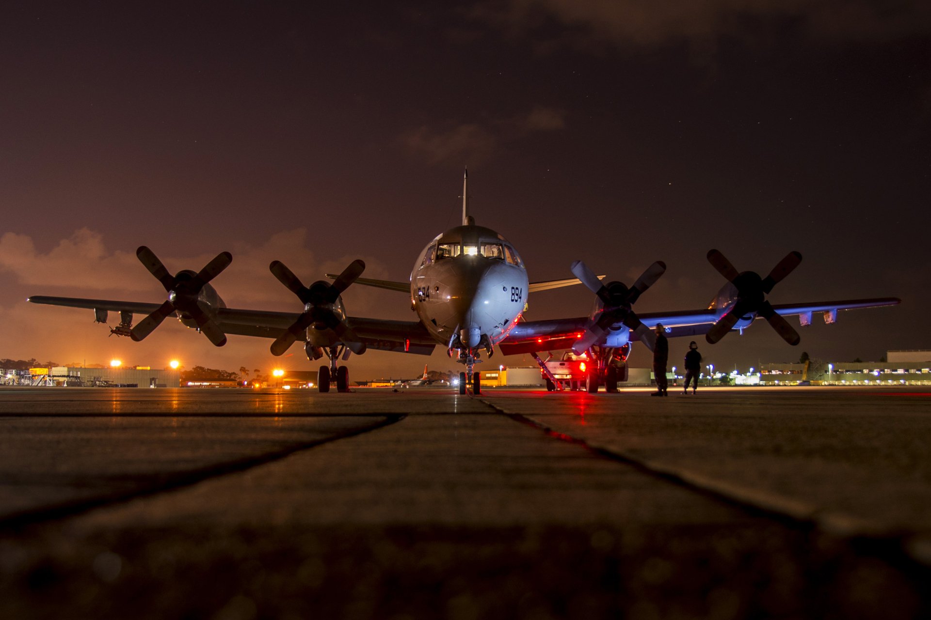A Lockheed P-3 Orion military aircraft is illuminated at night, showcasing its navy colors, with personnel and lights enhancing the scene on the tarmac. A stunning HD desktop wallpaper.