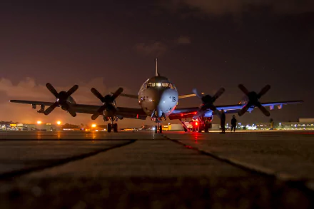 A Lockheed P-3 Orion military aircraft is illuminated at night, showcasing its navy colors, with personnel and lights enhancing the scene on the tarmac. A stunning HD desktop wallpaper.