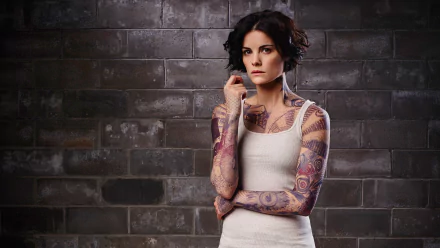 HD PC desktop wallpaper of a brunette with short hair and colorful full-sleeve tattoos in a tank top, posed against a brick wall — portrait of a TV-show character.