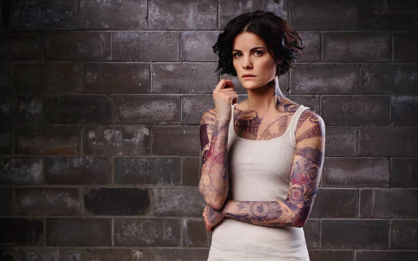HD PC desktop wallpaper of a brunette with short hair and colorful full-sleeve tattoos in a tank top, posed against a brick wall — portrait of a TV-show character.