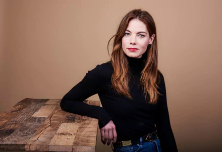 Studio portrait of a brunette woman wearing lipstick, seated by a rustic wooden table against a warm tan backdrop — high-resolution 8K Ultra HD desktop wallpaper.