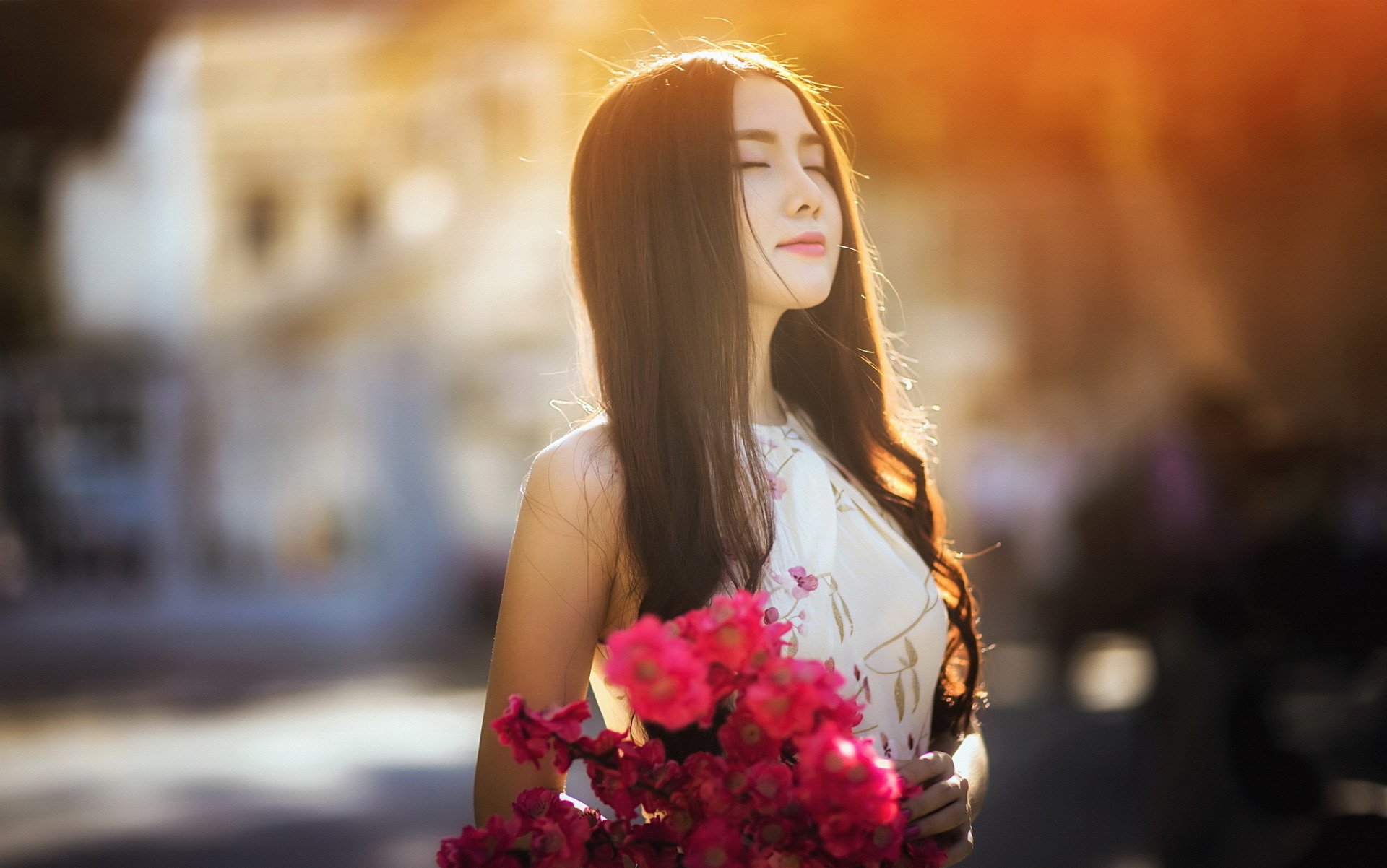 A serene Asian woman with long brunette hair and a white dress stands outdoors in the sunlight, holding vibrant pink flowers. The background is softly blurred, adding depth and a dreamy mood.