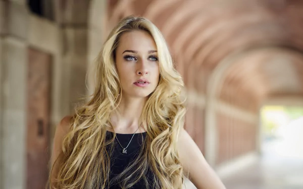 HD desktop wallpaper featuring a blonde woman with striking blue eyes, captured with a shallow depth of field highlighting her against a softly blurred architectural background.