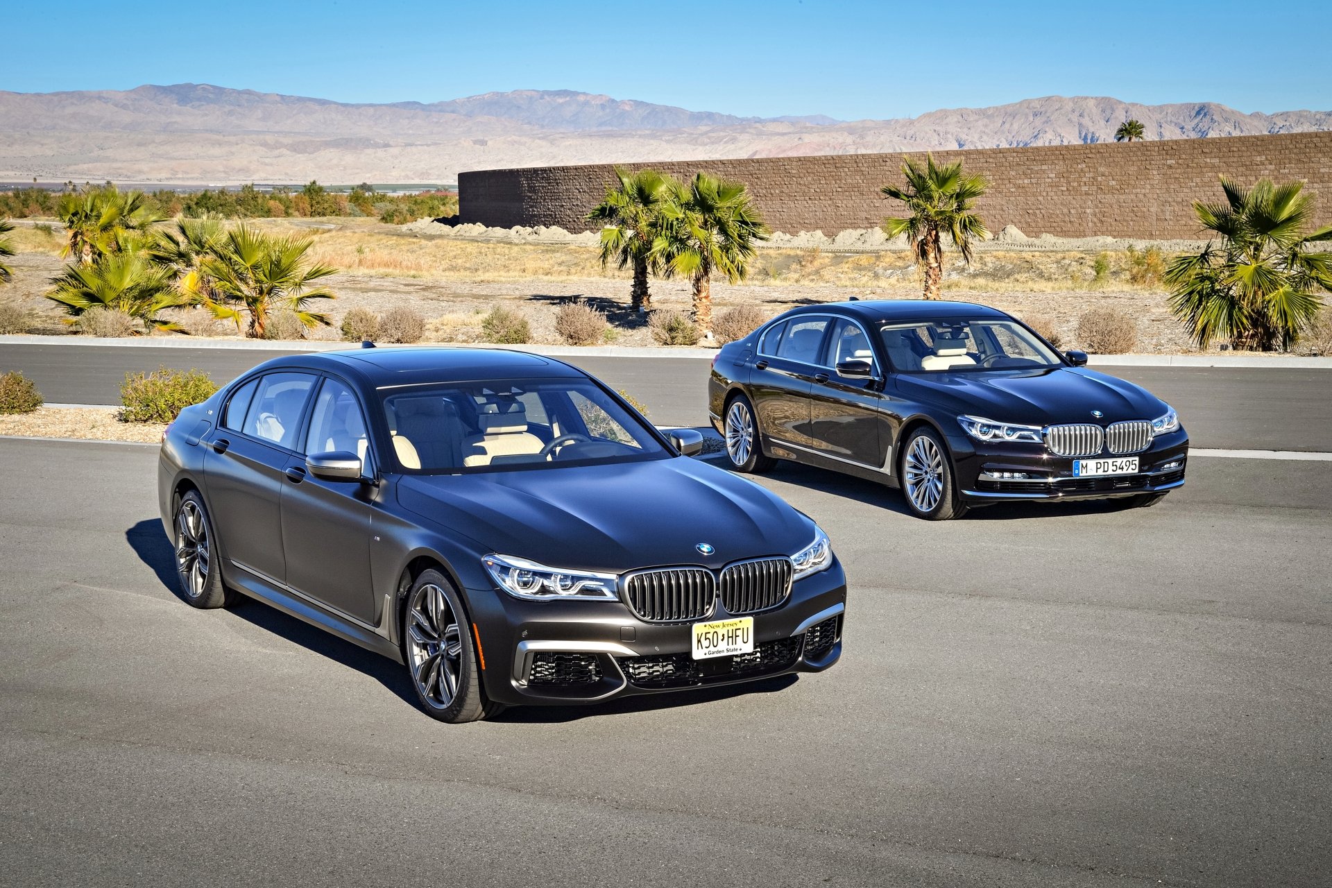 Two black BMW 7 Series vehicles parked on a desert road with palm trees and mountains in the background, captured in 4K Ultra HD desktop wallpaper quality.