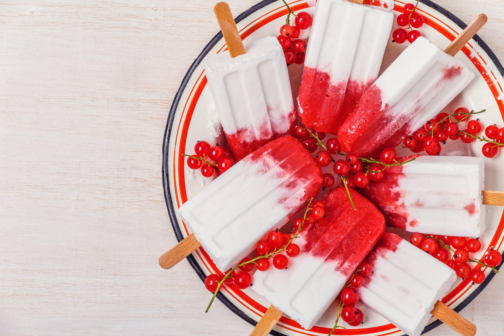 Bright 4K Ultra HD wallpaper showing creamy ice cream popsicles with red currants arranged on a white plate with red and blue stripes.