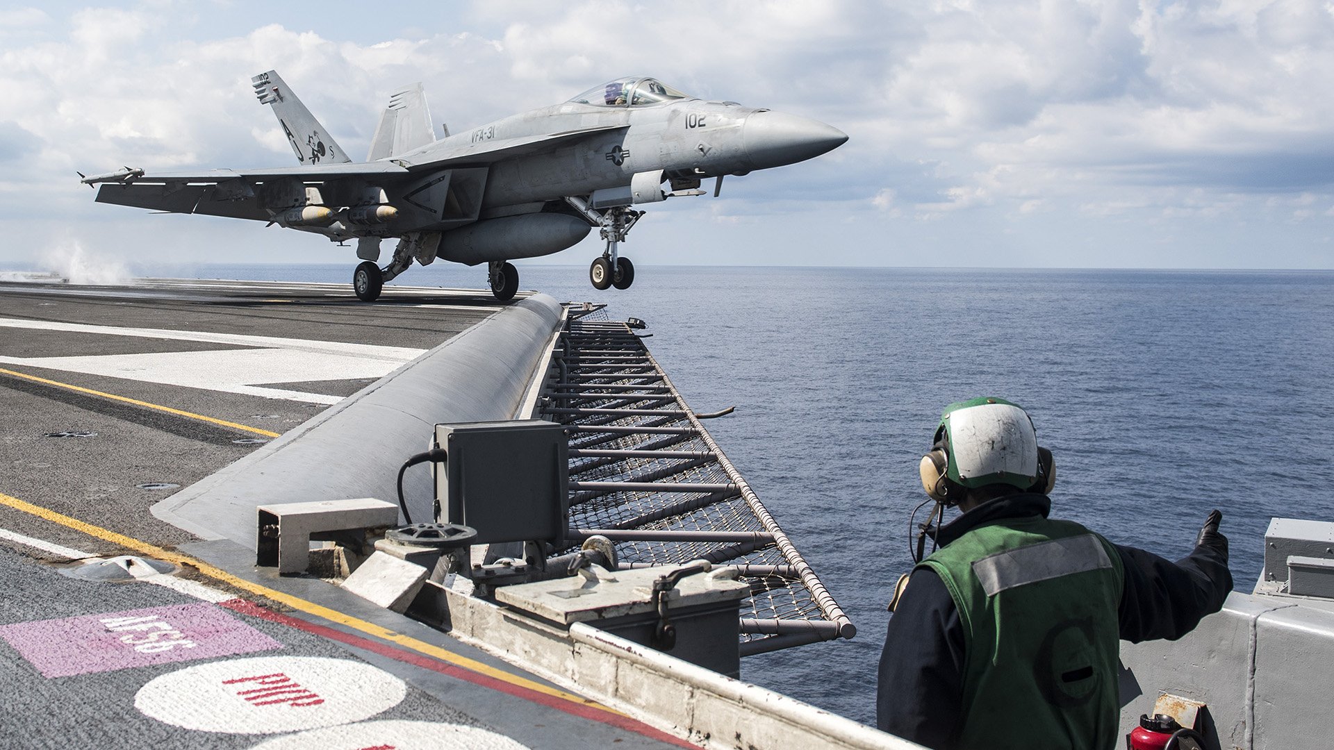 HD desktop wallpaper showing a Boeing F/A-18E/F Super Hornet warplane jet fighter landing on an aircraft carrier at sea under a cloudy sky.