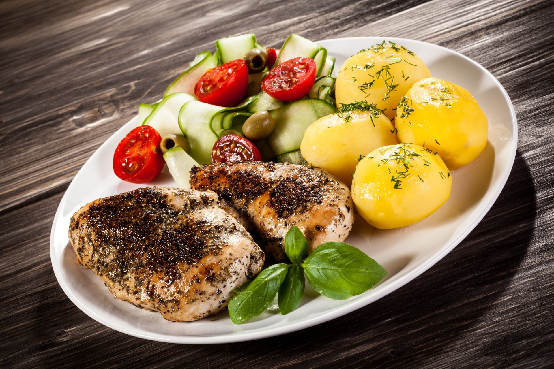 Plate of seasoned grilled meat served with boiled potatoes garnished with herbs and a side of fresh cucumber and cherry tomato salad, captured in 4K Ultra HD.