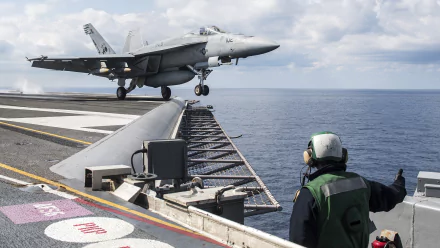 HD desktop wallpaper showing a Boeing F/A-18E/F Super Hornet warplane jet fighter landing on an aircraft carrier at sea under a cloudy sky.