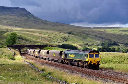A freight train travels through a lush, green countryside under a dramatic sky. This HD desktop wallpaper and background captures the beauty of a train journey through open landscapes.