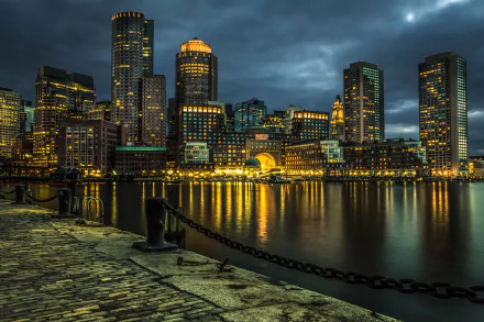 HD desktop wallpaper of Boston cityscape at night, featuring illuminated skyscrapers reflecting off the water, set against a dramatic, cloudy sky with a cobblestone path in the foreground.