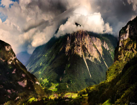 A dramatic Swiss mountain landscape with clouds and fog enveloping the peaks above a peaceful village nestled in the valley below.