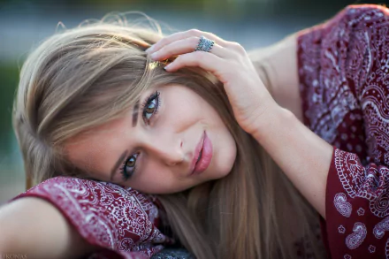 A high-definition desktop wallpaper featuring a blonde woman with hazel eyes and a captivating gaze, wearing a patterned maroon top, resting her head on her arm.