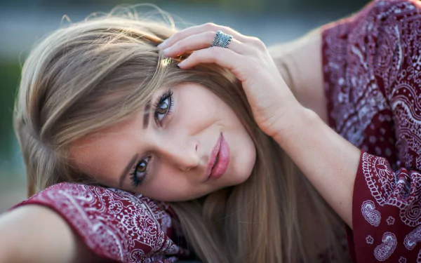 A high-definition desktop wallpaper featuring a blonde woman with hazel eyes and a captivating gaze, wearing a patterned maroon top, resting her head on her arm.