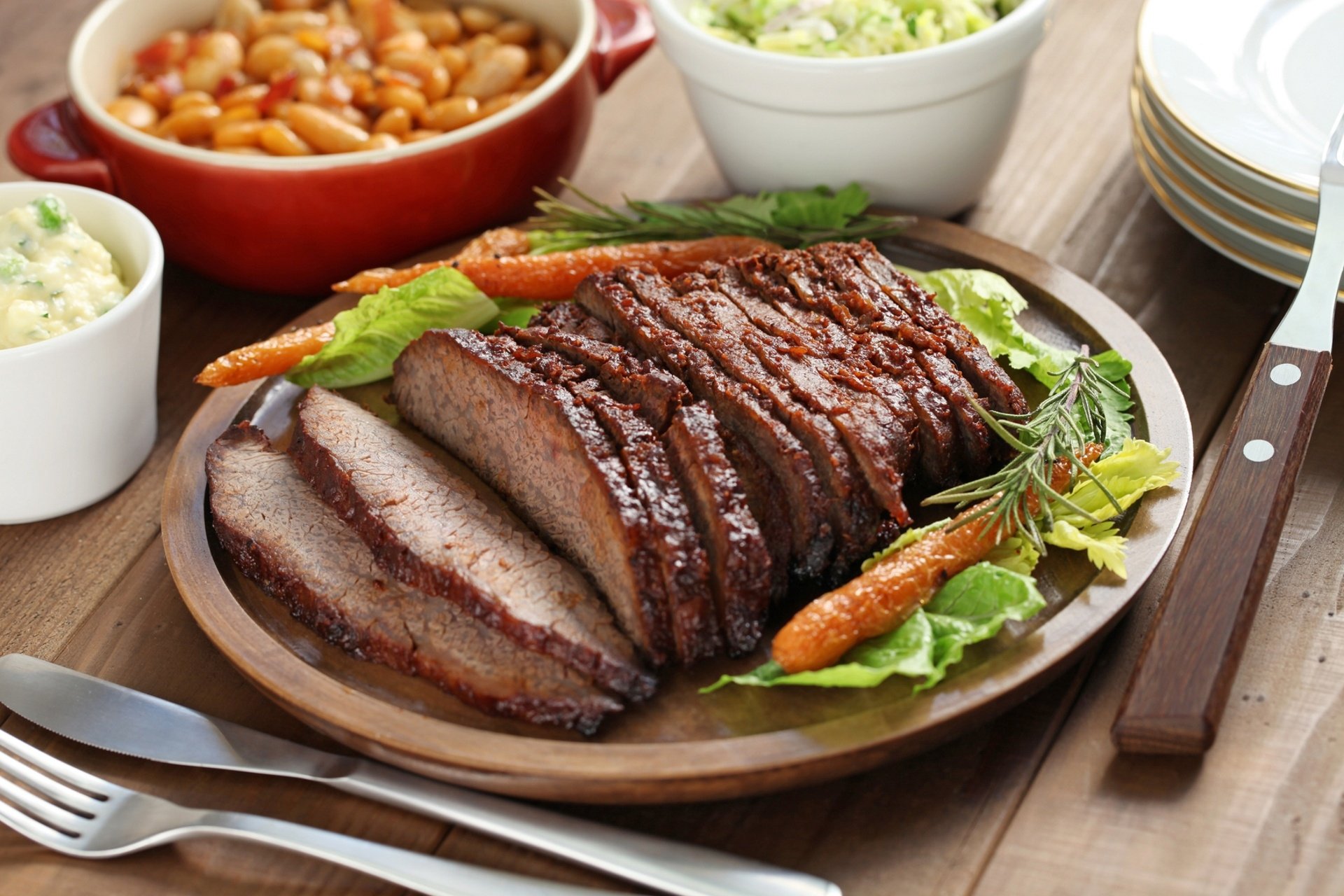 HD PC desktop wallpaper background of food: sliced roast meat brisket on a wooden platter with baby carrots, leafy greens and small bowls of side dishes.