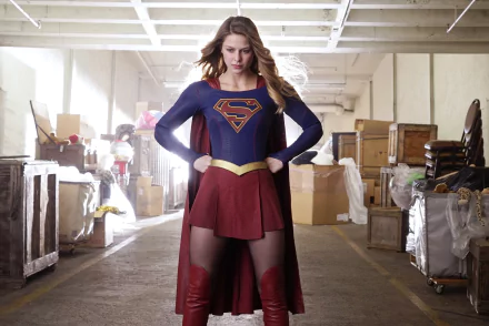 HD desktop wallpaper of Melissa Benoist as Supergirl from the TV show, wearing thigh boots, a cape, and a skirt, set in a warehouse. DC Comics superhero Supergirl stands confidently.