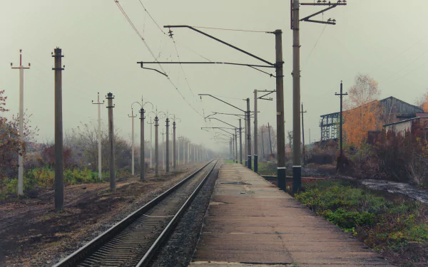 A 4K Ultra HD view of a deserted train station with railroad tracks and overhead power lines extending into the misty distance, surrounded by industrial structures.