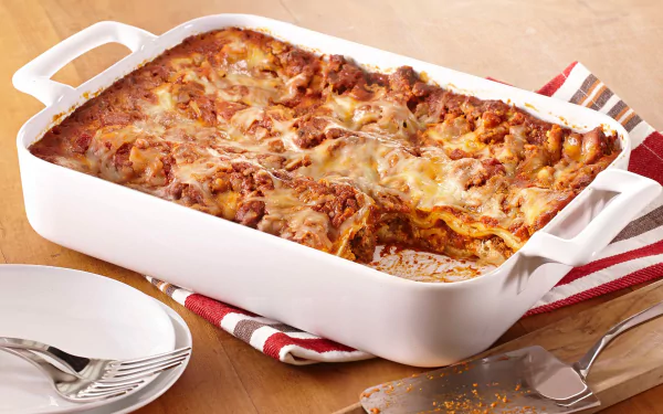 HD PC desktop wallpaper of a lasagna meal: baked, cheesy, browned in a white ceramic baking dish on a wooden table with plates and a spatula.