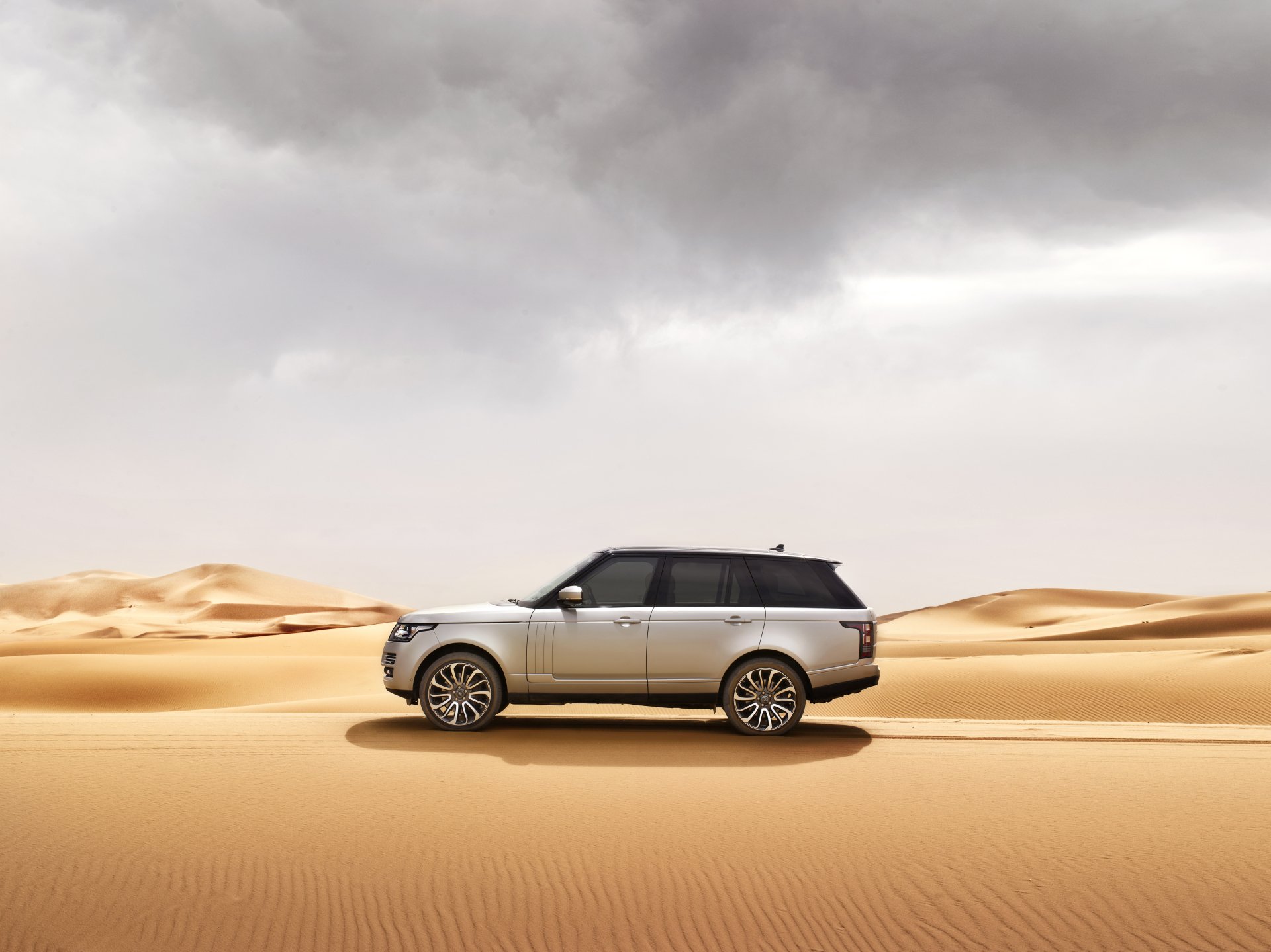Silver Range Rover SUV drives across sandy desert under a cloudy sky in this 4K Ultra HD PC desktop wallpaper and background.