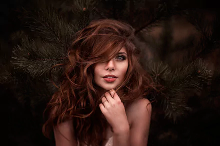 A striking redhead woman poses among lush green pine branches, her wavy hair cascading around her face. This HD desktop wallpaper showcases her model features in a captivating outdoor setting.