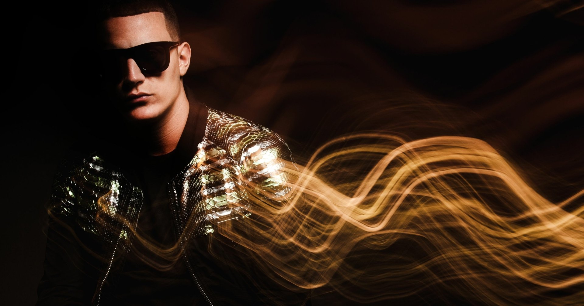 HD desktop wallpaper of a person with sunglasses and a shiny jacket, with golden light trails in the background, tagged with DJ Snake.