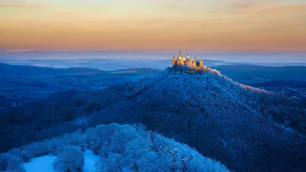 HD wallpaper featuring Hohenzollern Castle in Germany. The winter landscape showcases snow-covered hills and a stunning horizon.