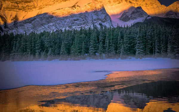 Winter scene of Two Jack Lake in Canada with icy water, a dense forest, and mountains glowing orange at sunset, captured in HD for a nature desktop wallpaper.