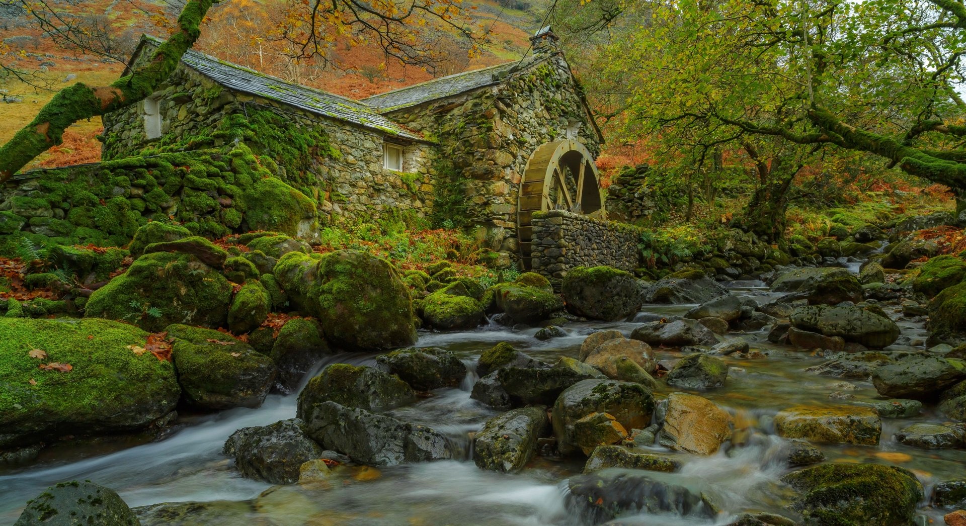 A serene HD desktop wallpaper featuring a moss-covered watermill set in a lush, green forest. A clear stream flows over stones in the foreground, adding to the tranquil scene.