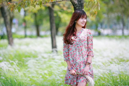 Asian brunette woman in a floral dress smiles softly in a sunlit field, with a shallow depth of field enhancing the serene natural background.